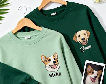 May include: Two crewneck sweatshirts, one in light green and the other in dark green, each featuring a custom embroidered dog portrait. The light green sweatshirt has a corgi design with the name "Vicky". The dark green sweatshirt has a labrador design with the name "Bruno".