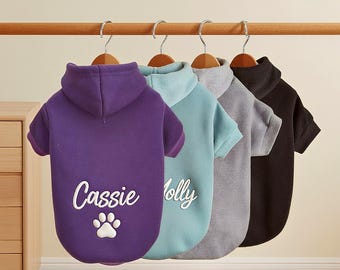 May include: Four dog hoodies in purple, teal, grey, and black hang on a wooden rail. The purple hoodie has a hood and the name "Cassie" embroidered with a paw print. The teal hoodie has the name "Molly". The hoodies have short sleeves.