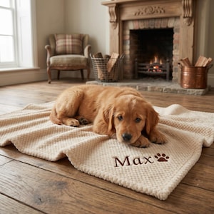 May include: A golden retriever puppy rests on a beige textured blanket with the name "Max" and a paw print embroidered in brown. The blanket is on a wooden floor, with a fireplace and chair in the background.