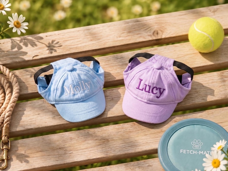 May include: Two personalized dog baseball caps, one light blue with "Molly" embroidered, the other lavender with "Lucy". A tennis ball and a "Fetch-Mate" frisbee are also visible, suggesting a pet-themed product.
