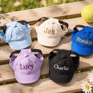 May include: A selection of dog baseball caps in light blue, beige, blue, purple, and black. Each cap is embroidered with a name. A tennis ball and daisies are also visible, arranged on a wooden surface.