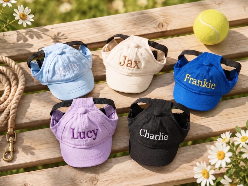 May include: A collection of personalized dog baseball caps in various colors: light blue, beige, blue, purple, and black. Each cap has a name embroidered on the front. A tennis ball and daisies are also visible.