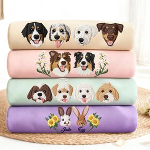Puede incluir: Cuatro almohadas bordadas apiladas, en beige, rosa, verde menta y morado. Cada almohada presenta caras de animales detalladas: perros y conejos. La almohada inferior tiene los nombres "Jude" y "Egg" bordados.