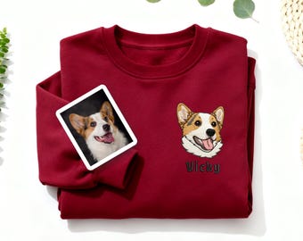 May include: A burgundy sweatshirt featuring an embroidered corgi dog face and the name "Vicky" in black script. A small photo of a corgi is attached to the sweatshirt. The sweatshirt is made of a soft material.