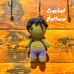 May include: Green crocheted Hulk character with purple pants and black hair. The character is attached to a silver key ring. The text "Crochet Pattern" is visible in the background.