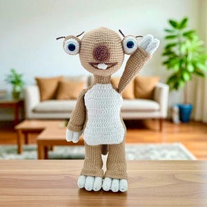 Könnte beinhalten: Handgefertigte Häkelpuppe in Form einer Zeichentrickfigur. Die Puppe hat einen beigefarbenen Kopf, Arme und Beine, große blaue Augen und einen weißen Bauch. Sie winkt mit einem Arm. Die Puppe steht auf einer Holzoberfläche.