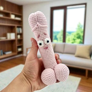 May include: A light pink, crocheted phallus with a smiling face and large, round testicles. The handmade item has white eyes and a smiling mouth. The background includes a living room with a couch, rug, and bookshelves.
