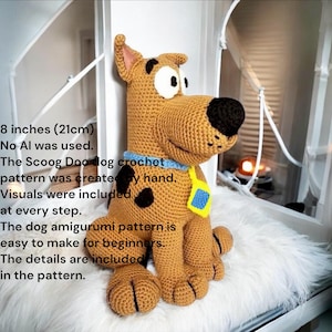 May include: A crocheted Scooby Doo dog amigurumi, about 21cm tall. The dog is brown with black spots, a black nose, and a blue collar with a yellow tag. The image includes text stating the pattern is handmade and easy for beginners.