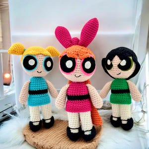 Puede incluir: Tres muñecas de ganchillo hechas a mano, cada una representando un personaje de Las Supernenas. Las muñecas presentan distintos colores de pelo: amarillo, naranja y negro. Visten vestidos en azul, rosa y verde, con detalles en negro. Cada muñeca tiene ojos grandes y una cara sonriente.