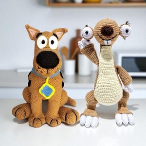 May include: Two crocheted character dolls. One is a brown dog with a black nose and a blue collar with a yellow and blue tag. The other is a brown sloth with large eyes and a cream-colored belly. Both are handmade.