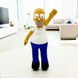 Könnte beinhalten: Handgefertigte Häkelpuppe von Homer Simpson. Die Puppe hat einen gelben Kopf, Arme und Hände, ein weißes Hemd, blaue Hosen und schwarze Schuhe. Ein Arm ist zum Winken erhoben.