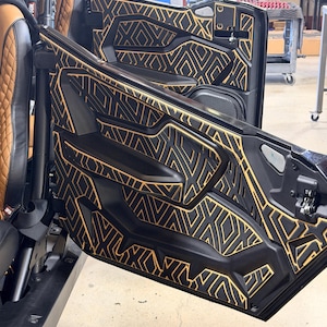 Pode incluir: Painéis de porta de veículo pretos com um padrão geométrico dourado. Os painéis estão abertos, revelando um assento castanho e preto. Os painéis da porta têm um design complexo e repetitivo.