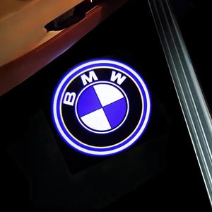 Puede incluir: Logotipo BMW iluminado, con el emblemático emblema azul y blanco, sobre un fondo oscuro. El logotipo está rodeado por una luz blanca y azul brillante, lo que mejora su visibilidad. La imagen sugiere un accesorio automotriz.