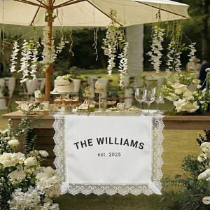 Puede incluir: Una mesa decorada para una celebración, con un camino de mesa de encaje blanco con el texto "THE WILLIAMS est. 2025". La mesa está adornada con una tarta, magdalenas y arreglos florales. Una sombrilla abierta proporciona sombra.