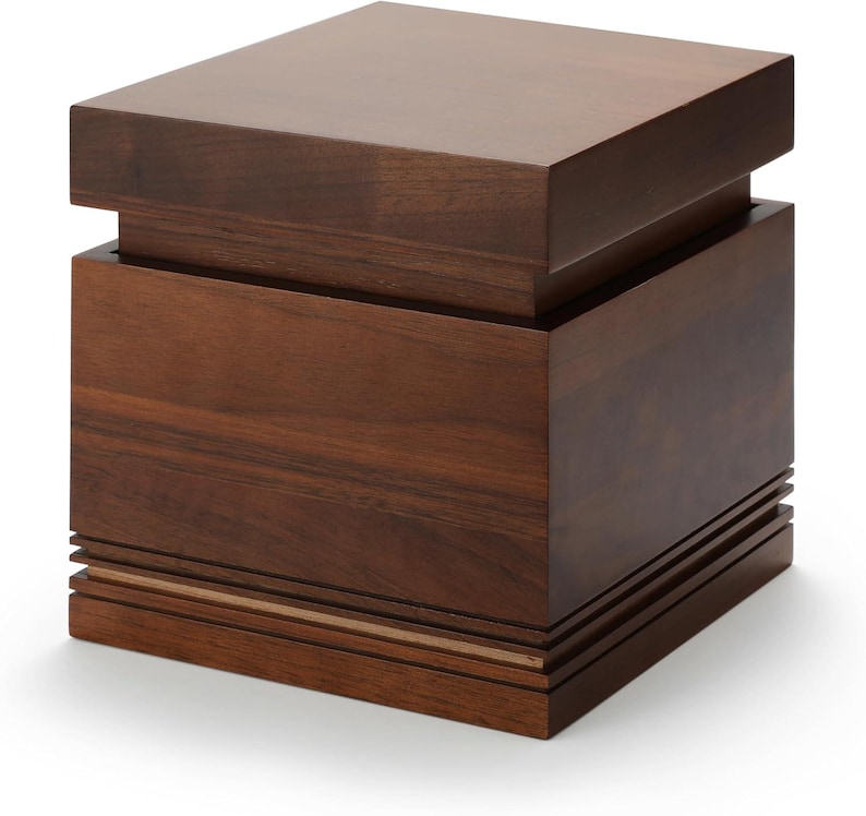 May include: A dark brown wooden box with a square shape. The box has a layered design, with a top section that overhangs the main body. The base features horizontal grooves and a thin, lighter-colored accent.