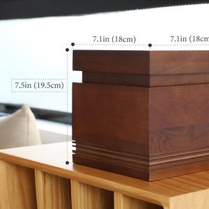 May include: A dark brown wooden box, 7.1 inches (18cm) wide and 7.5 inches (19.5cm) tall, with a layered design. The box is sitting on a light brown wooden surface. The box has a rectangular shape and is likely used for storage or display.