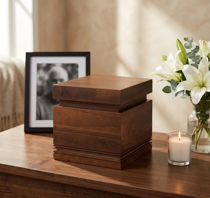 May include: A dark brown wooden urn with a square shape and a layered design. A framed black and white photo and a lit candle in a glass are also on the wooden surface. White lilies in a glass vase are in the background.