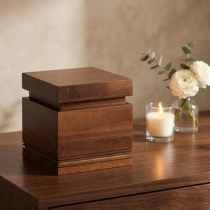 May include: A dark brown wooden box with a stepped design. A lit candle in a glass and a small vase of white flowers are next to the box. The box is on a dark brown wooden surface.