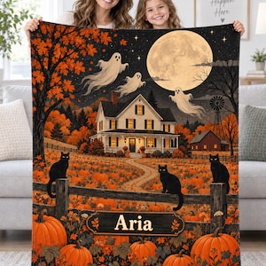 May include: A rectangular throw blanket featuring a Halloween scene with a white house, a large full moon, three ghosts, and three black cats sitting on a wooden fence. The foreground displays orange pumpkins and the name Aria on a wooden sign.