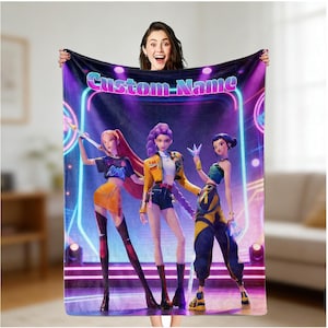 Puede incluir: Una manta de felpa con un gráfico vibrante de tres personajes femeninos animados con atuendos elegantes. La manta tiene un fondo iluminado con neón con el texto "Custom Name". La manta es sostenida por una persona.