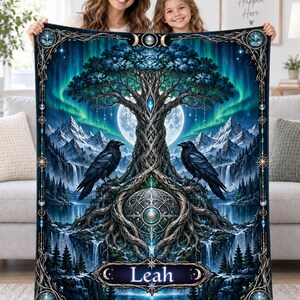 Puede incluir: Una manta rectangular con un árbol central de raíces intrincadas, dos cuervos negros, una luna llena, montañas nevadas y una cascada. El nombre Leah aparece impreso en una banda decorativa en la parte inferior, enmarcado por motivos celestiales.