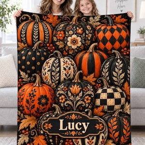 May include: A rectangular fleece blanket featuring a pattern of various pumpkins in black and orange. The pumpkins display diverse designs including stripes, polka dots, checkered patterns, and floral motifs. A central black plaque displays the name Lucy.