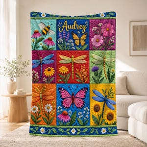 Puede incluir: Una manta acolchada con una cuadrícula de nueve paneles coloridos que muestran ilustraciones de abejas, mariposas y libélulas entre varias flores. El panel central superior muestra el nombre Audrey en texto cursivo amarillo.