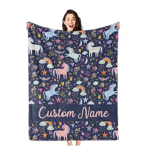 Könnte beinhalten: Eine marineblaue Decke mit einem sich wiederholenden Muster aus Einhörnern, Regenbogen, Sternen und Blumen. Die Decke hat einen Abschnitt mit dem Text "Custom Name" in rosa Schrift. Die Decke wird von einer Person gehalten.