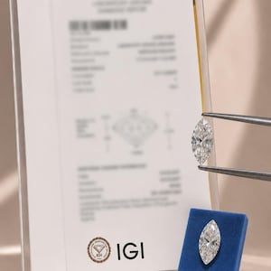 Könnte beinhalten: Ein Diamant im Marquise-Schliff, gehalten von einer Metallpinzette, neben einem zweiten identischen Diamanten auf einem kleinen blauen quadratischen Sockel, begleitet von einem vertikalen IGI-Laborbericht in einer transparenten Hülle.