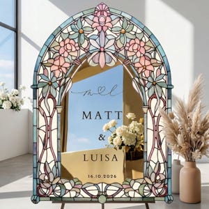 May include: A stained glass wedding sign with a floral design in pastel shades. The sign displays the names "Matt & Luisa" and the date "16.10.2026". It is set on an easel, with floral arrangements and pampas grass in the background.