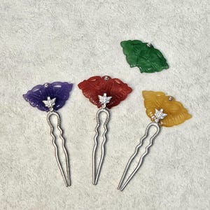 May include: Four colorful hair pins with flower-shaped tops. The pins are purple, red, yellow, and green. Each pin has a silver stem and a small, decorative accent. The hair accessories are set against a neutral background.