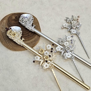 May include: Three decorative hair sticks with floral and pearl accents. One is gold-toned, the other two are silver-toned. The hair sticks feature intricate designs with white, gold, and burgundy accents. The hair accessories are displayed on a wooden surface.