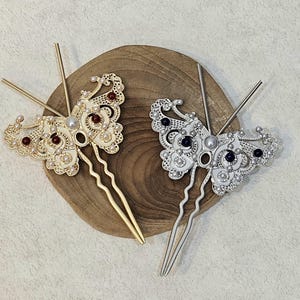 May include: Two decorative butterfly hair pins. One is gold-toned with red and pearl accents, the other is silver-toned with dark purple and pearl accents. Both pins have long, slender prongs.