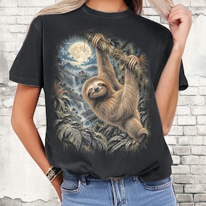 Könnte beinhalten: Ein graphitfarbenes T-Shirt mit einem Grafikdruck eines braunen Faultiers, das an einem Ast in einer Dschungellandschaft hängt, mit Vollmond und Wasserfällen im Hintergrund. Das Wort GRAPHITE ist oben links im Bild aufgedruckt.