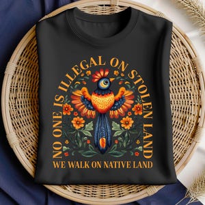 May include: Black t-shirt with a colorful floral design and a bird in the center. The text "No one is illegal on stolen land. We walk on native land" is printed in orange on the shirt.