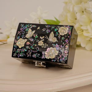 Puede incluir: Un joyero rectangular negro con un intrincado diseño de nácar incrustado en la tapa. La decoración muestra mariposas iridiscentes y patrones florales con acentos brillantes en verde, rosa y blanco, junto con un cierre de metal.