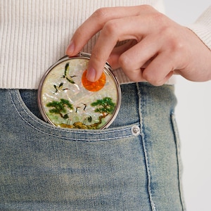 Pode incluir: Um espelho compacto redondo e prateado com uma cena detalhada e colorida de guindastes, árvores e um sol. O espelho está sendo segurado em um bolso de jeans azul. A obra de arte é definida contra um fundo perolado.