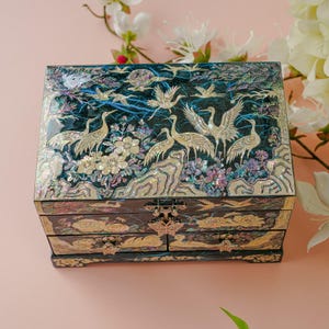 May include: A black jewelry box with a detailed mother-of-pearl inlay featuring cranes, flowers, and foliage. The box has multiple drawers and a hinged lid. The color palette includes black, white, and iridescent hues.