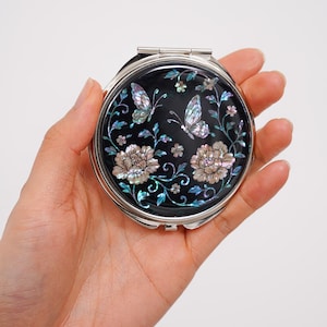 Pode incluir: Um espelho compacto redondo de metal prateado com um embutido preto decorado com motivos florais e de borboletas em madrepérola iridescente. O design inclui duas grandes flores desabrochando, vinhas delicadas e duas borboletas.