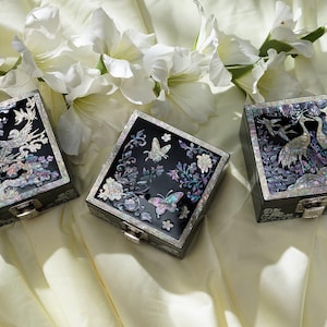 May include: Three square black lacquer boxes with hinged metal latches. Each lid features intricate iridescent mother-of-pearl inlay designs, including depictions of birds, butterflies, and floral patterns with shimmering purple and blue tones.