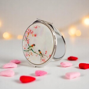 May include: A silver compact mirror with a decorative design featuring two birds perched on a flowering branch. The mirror is open, revealing a reflective surface. Pink and red decorative shapes are scattered around the mirror.
