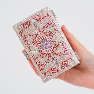 Peut inclure: Une boîte rectangulaire ornée avec un fond rouge et une incrustation de nacre complexe. Le motif présente des motifs floraux et d'oiseaux, avec une fleur centrale et deux oiseaux. La boîte est tenue dans une main et possède des fermoirs en métal argenté.