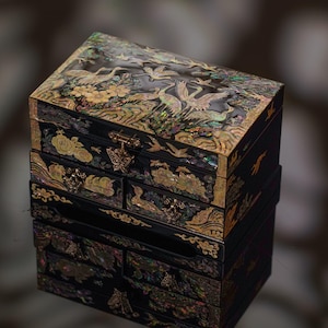 May include: Black jewelry box with multiple drawers, adorned with iridescent mother-of-pearl inlay. The box features detailed depictions of cranes, flowers, and other decorative elements. A butterfly-shaped clasp secures the top. The box is reflected in the surface below.