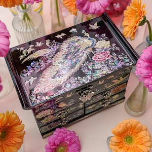 May include: A dark purple jewelry box featuring an intricate mother-of-pearl inlay of a peacock and floral patterns on the lid. The rectangular chest includes multiple drawers with butterfly-shaped metal pulls and a glossy, reflective finish.
