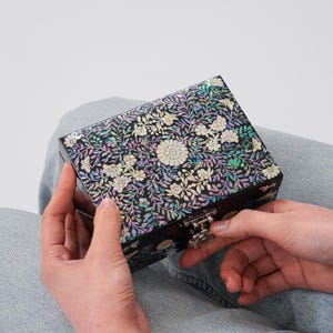 May include: A rectangular black jewelry box featuring an intricate inlaid mother-of-pearl floral and vine pattern across the lid and front. The box includes a silver-toned metal latch mechanism on the front for securing the lid closed.