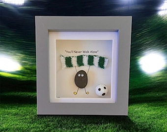 Puede incluir: Obra de arte enmarcada con una figura de guijarro sosteniendo una bufanda verde y blanca, con un balón de fútbol. El texto "You'll Never Walk Alone" está encima de la bufanda. El marco es gris, sobre un fondo verde.