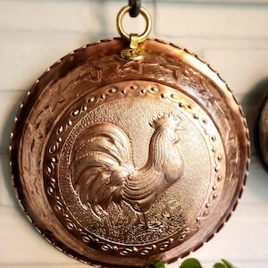 May include: A copper-colored, round wall hanging with a detailed embossed rooster design. The decorative plate has a scalloped edge and is suspended by a brass ring and hook. The plate is approximately 12 inches in diameter.