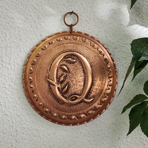 Könnte beinhalten: Kupferfarbene Metall-Wanddekoration mit dem Buchstaben „Q“ und einem Olivenzweig-Design. Die runde Plakette hat einen dekorativen Perlenrand und einen kleinen Ring zum Aufhängen. Ein einzigartiges Stück für die Inneneinrichtung.