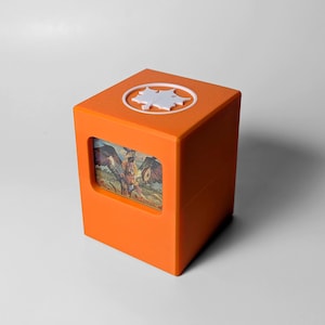 May include: An orange cube-shaped box with a white leaf-shaped design on top. A rectangular window on the front displays a colorful illustration of a mythical creature with wings. The box is made of a solid, opaque material.