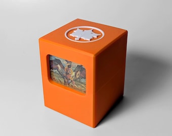 May include: An orange cube-shaped box with a white leaf-shaped design on top. A rectangular window on the front displays a colorful illustration of a mythical creature with wings. The box is made of a solid, opaque material.